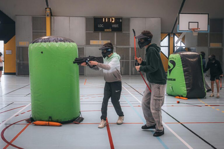 Two people playing indoor paintball wearing masks and using inflatable barriers for cover.